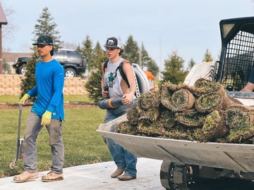 Two workers preparing landscaping with rolled sod and equipment outdoors.