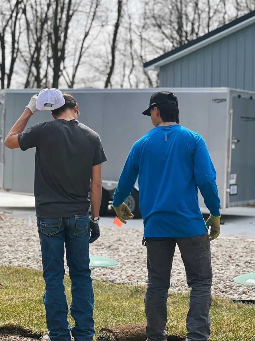 Two men standing outside near gravel and grass, viewed from behind.