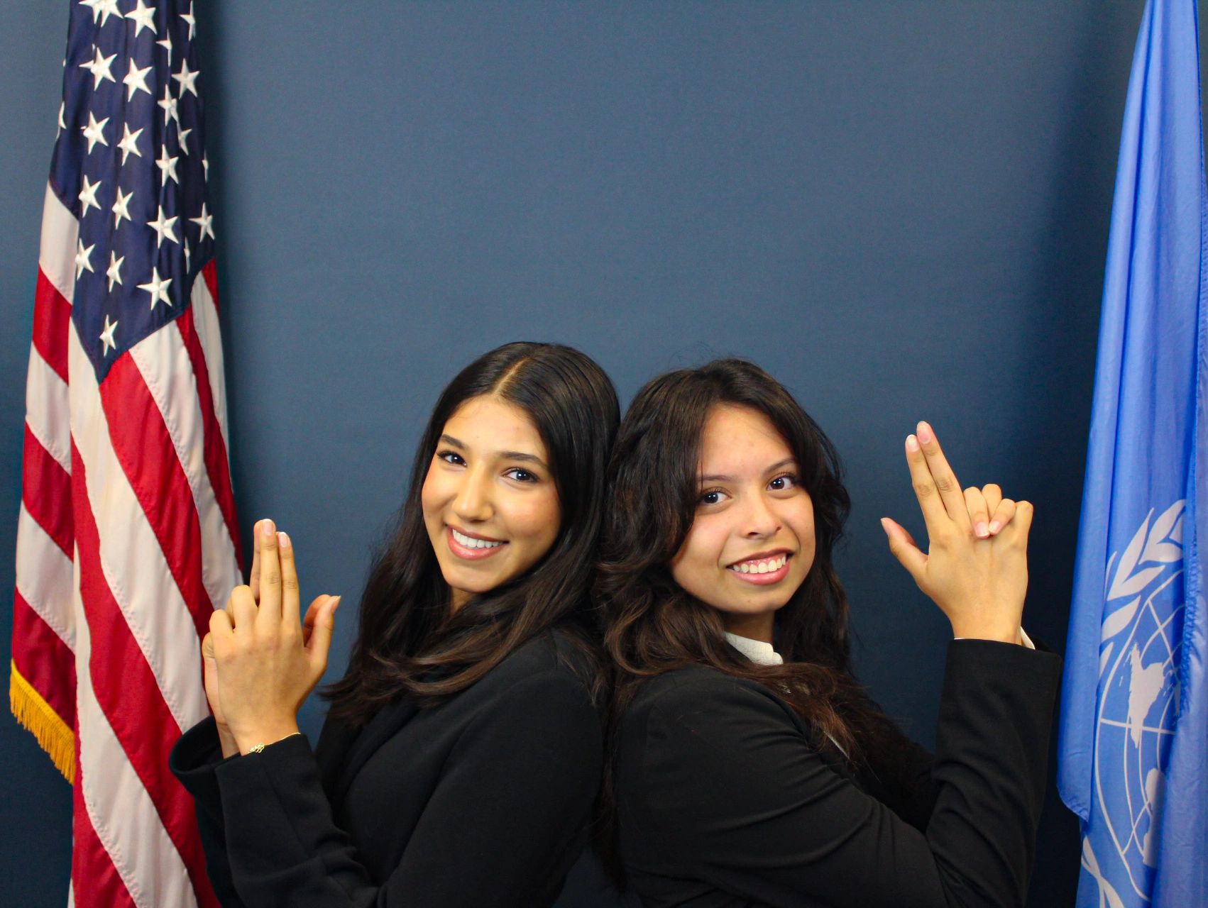 Two women smiling, posing back-to-back with finger guns, flags in background.