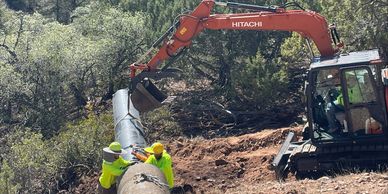Workers install a large pipeline in a forested area using heavy machinery.
