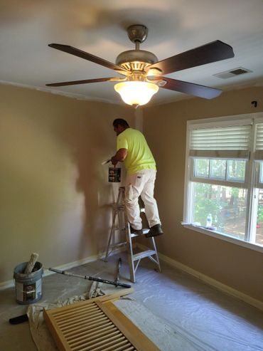 A man painting a beige wall in a room with a ceiling fan and window.