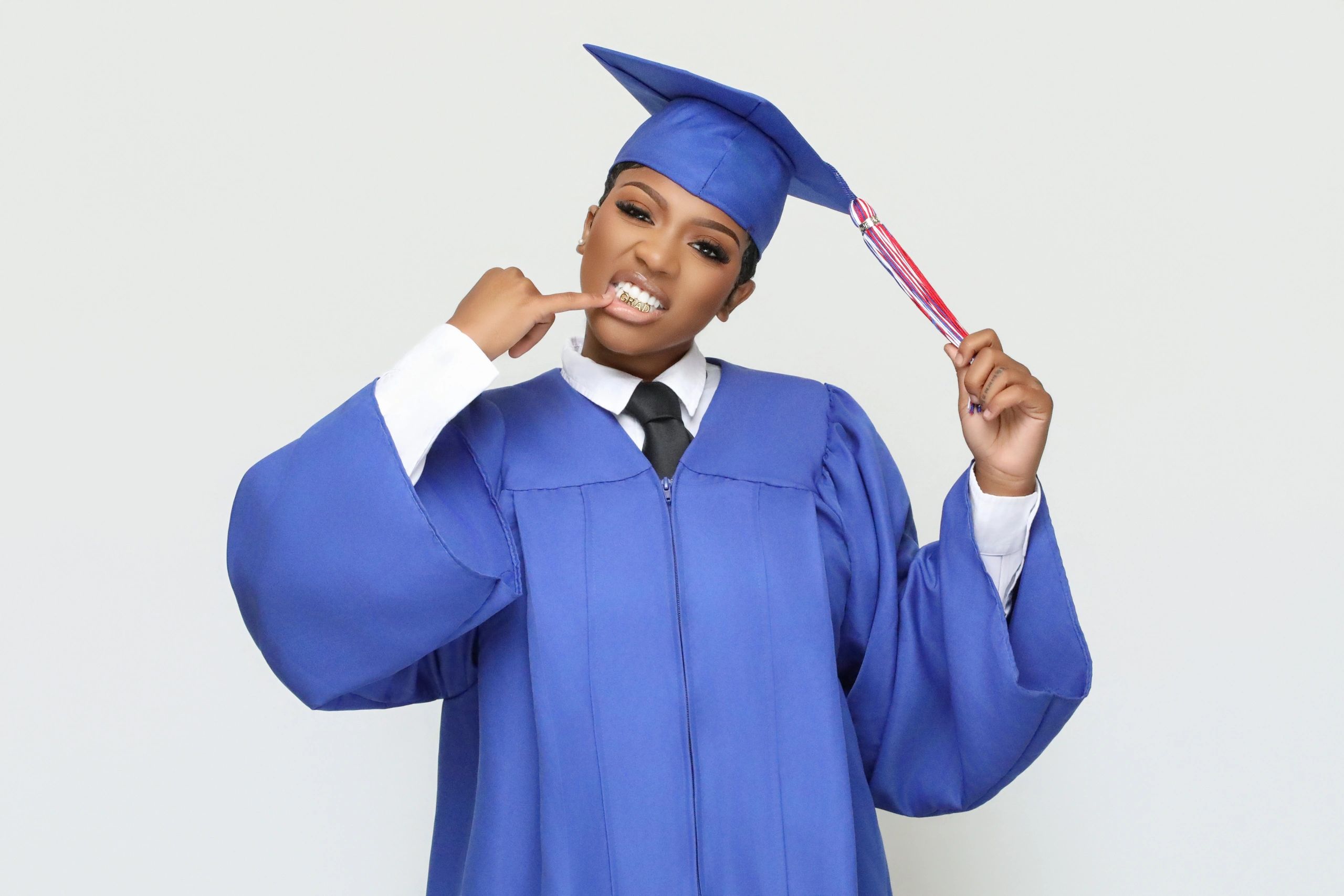 Graduate in blue cap and gown showing off her grill with 'GRAD' letters.