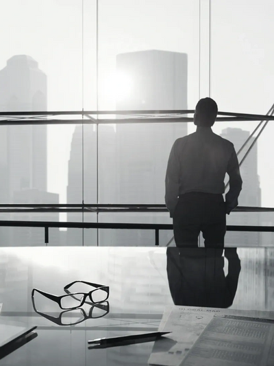A man stands by a large window overlooking a city skyline, with glasses and papers on a reflective desk.