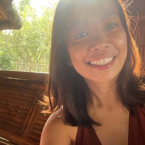 Young woman smiling with sunlight and greenery in the background.