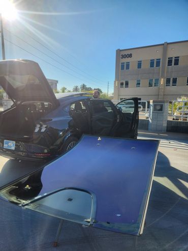 A person works on a black car with its doors and trunk open under bright sunlight.