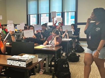 Students in a classroom hold up papers while a person stands and observes.