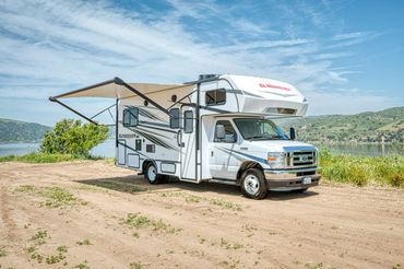 A white RV with an extended awning parked near a lake on a sunny day.
