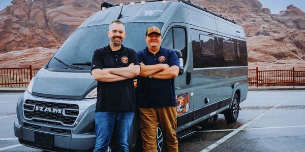 Two men standing confidently in front of a RAM van in a rocky outdoor setting.