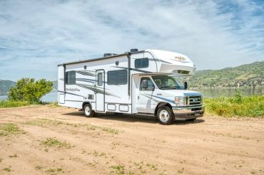 White motorhome parked on dirt road near water and hills.
