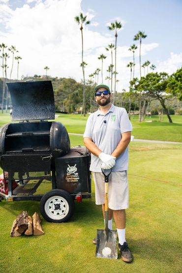 Man with shovel on golf course near barbecue smoker.