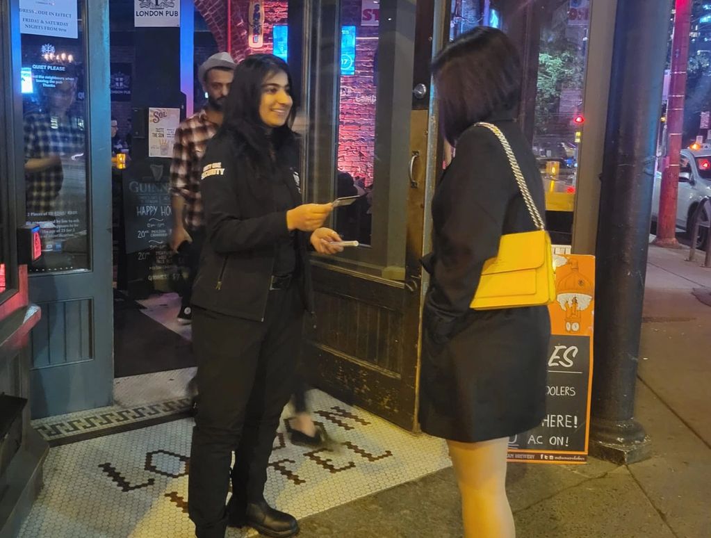 Security staff checks ID of a woman at a pub entrance at night.
