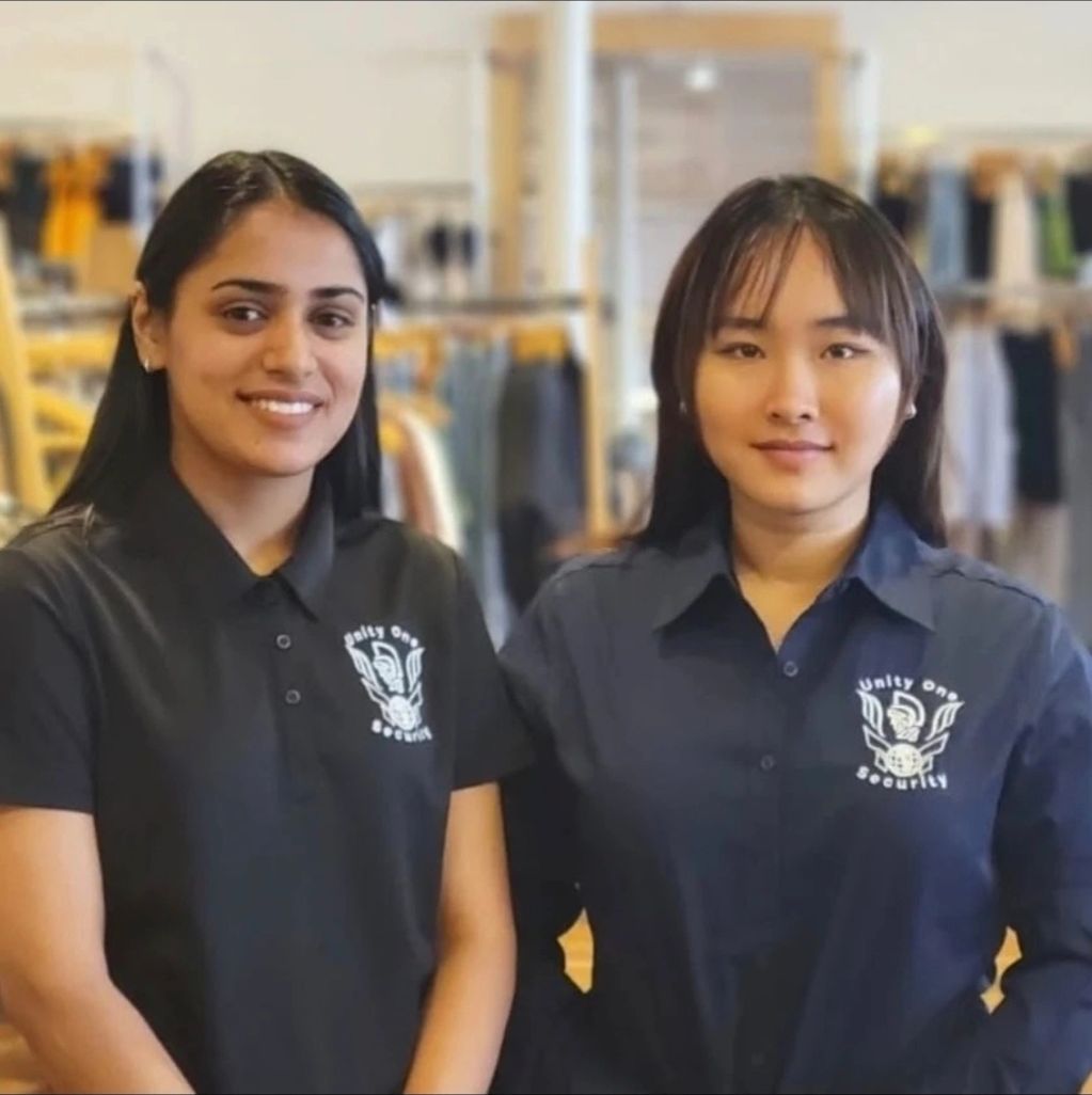 Two women wearing Unity One Security uniforms posing indoors.