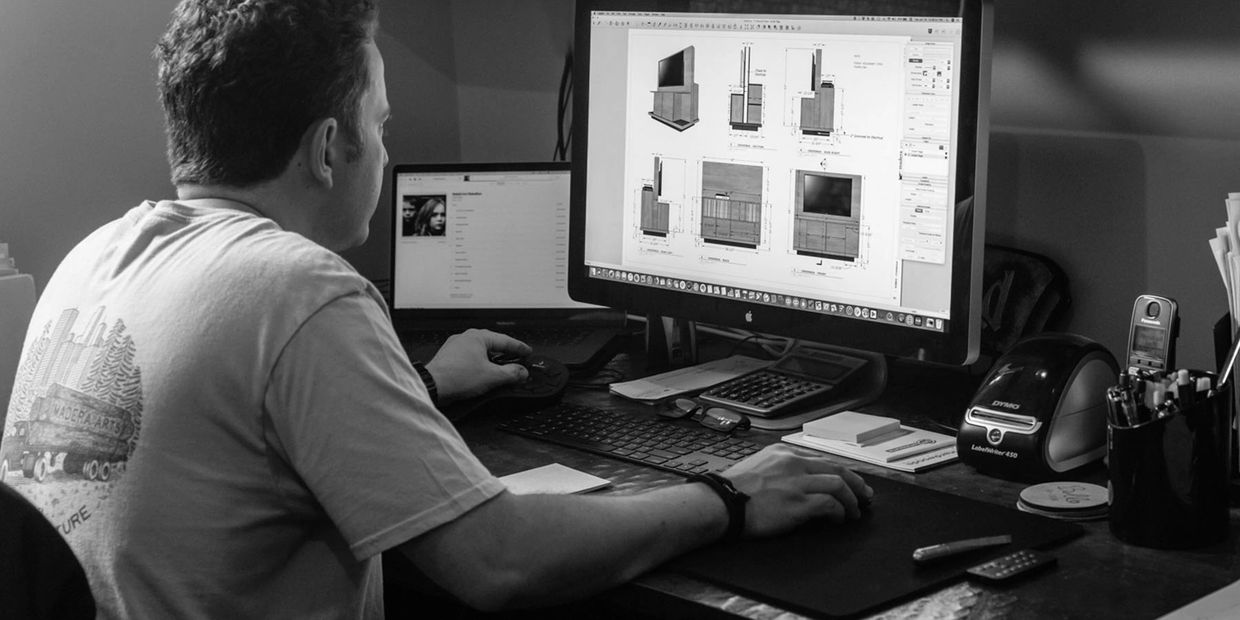 A man working on furniture design plans on a desktop computer at a wooden desk.