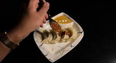 Hand drizzling spicy sauce over steamed dumplings on a marble-patterned plate.