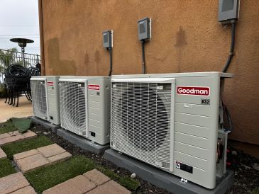 Three Goodman air conditioning units installed outside a house with a brown wall.