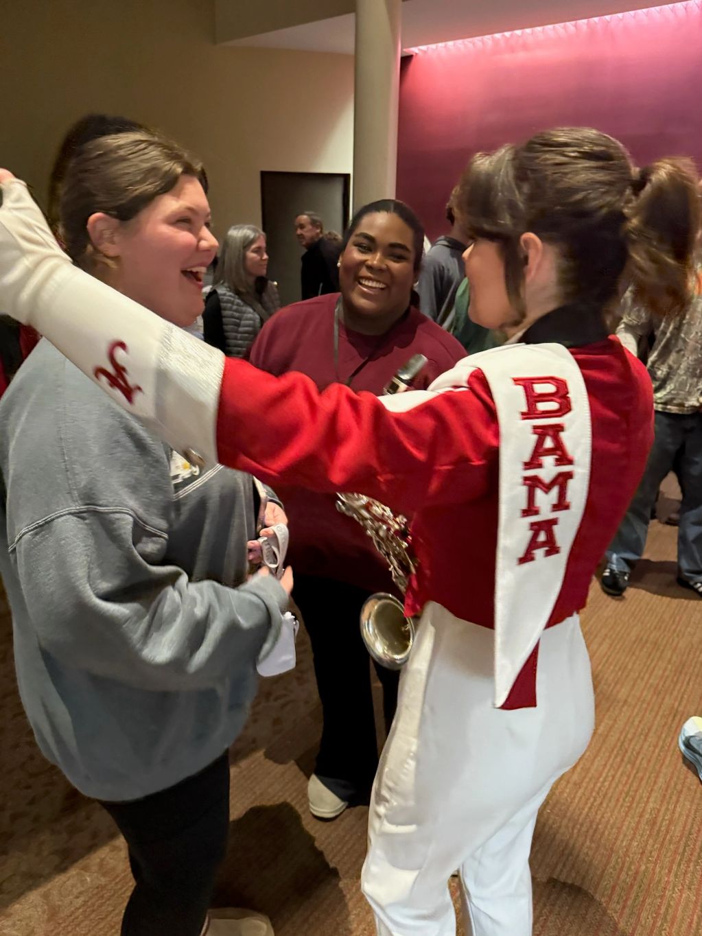Three women happily interacting indoors, one in a BAMA band uniform holding a saxophone.