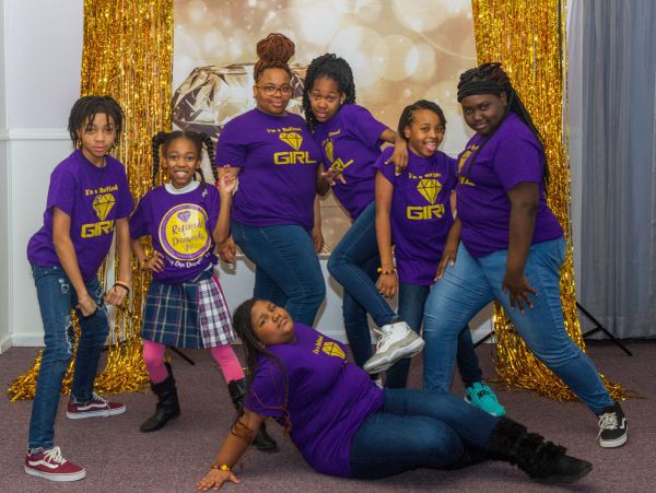 Group of girls in purple shirts posing in front of a golden backdrop.