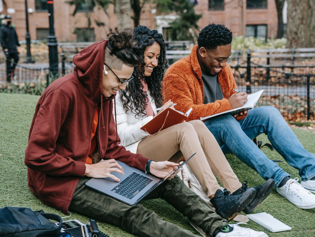 Three friends studying together outdoors, smiling and writing.
