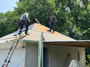 Two workers repairing a garage roof surrounded by trees on a sunny day.