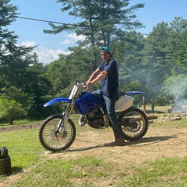 Person standing beside a blue dirt bike outdoors.