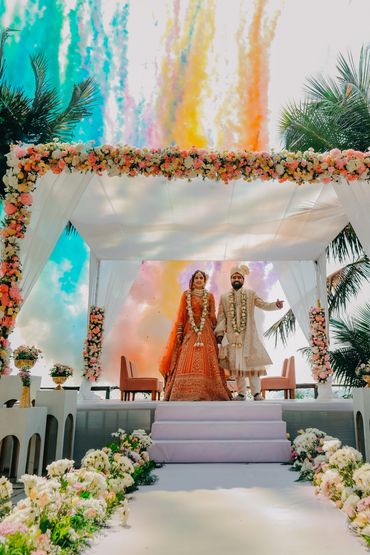 A bride and groom stand under a flower-adorned canopy with colorful smoke in the background.