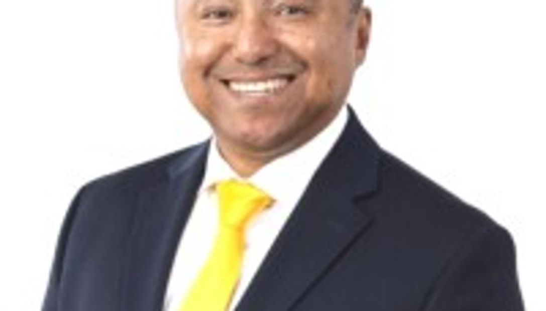 Smiling man wearing a dark suit and bright yellow tie against a white background.