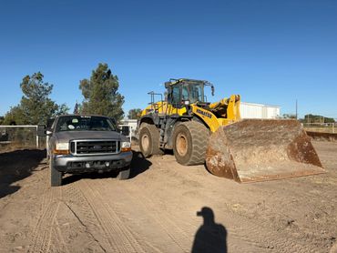 A silver Ford truck parked next to a yellow Komatsu bulldozer on a dirt lot under a clear blue sky.