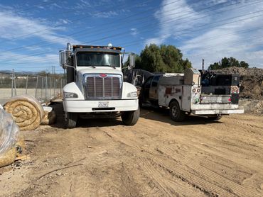 White Peterbilt truck and utility pickup truck parked on dirt road under blue sky.