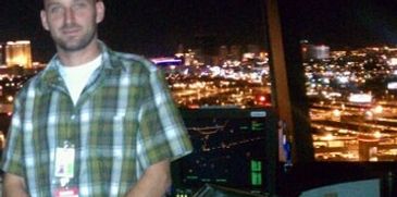 Man in a plaid shirt standing indoors with city lights visible through the window behind him at night.