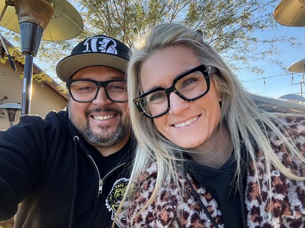 A smiling couple wearing glasses enjoys a sunny outdoor setting.