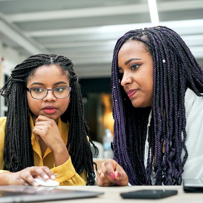 Two women with braided hair focused on a laptop screen in a workspace.