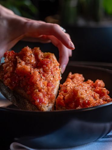 Hand placing tomato bruschetta on toasted bread in a black bowl