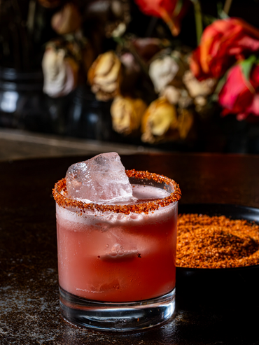 Pink cocktail with chili-salt rim and ice on a dark bar, blurred flowers behind