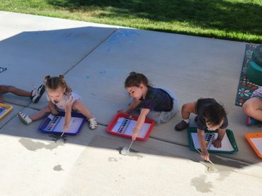 Five children painting on pavement with ice and watercolors outdoors on a sunny day.