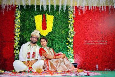 Couple sitting in traditional attire at a colorful floral wedding backdrop.