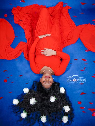 Pregnant woman in a red dress lying on blue with white flowers in her hair.