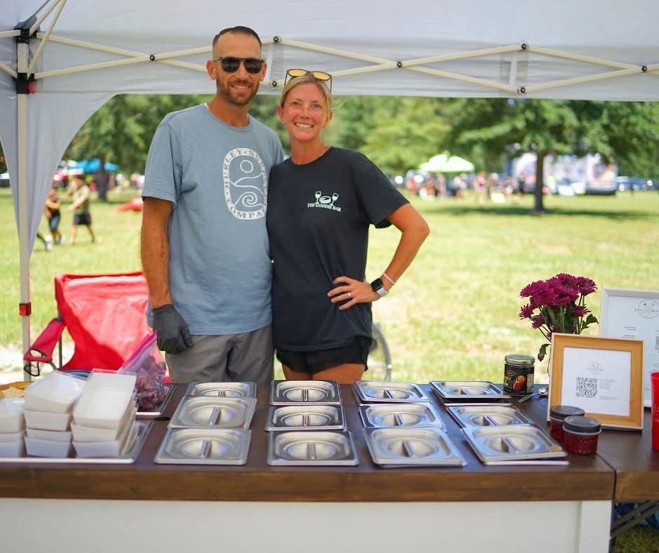 Two vendors standing behind a charcuterie cart with covered trays at an outdoor event.