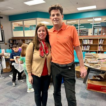 Two adults posing together in a library with children and bookshelves in the background.