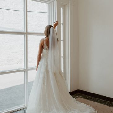 Bride in white gown standing by a large arched window.