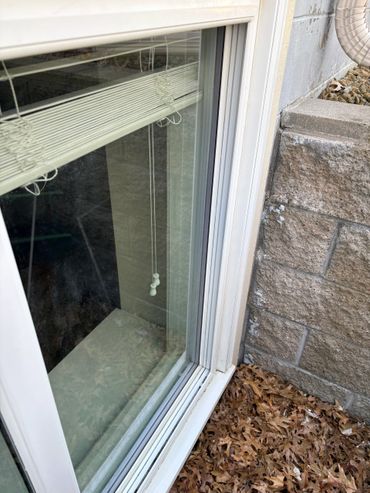 Close-up of a basement window with blinds and fallen leaves outside.