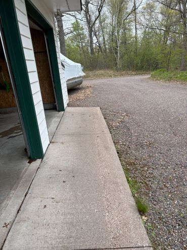 Open garage door with a covered boat beside a gravel road and forest.