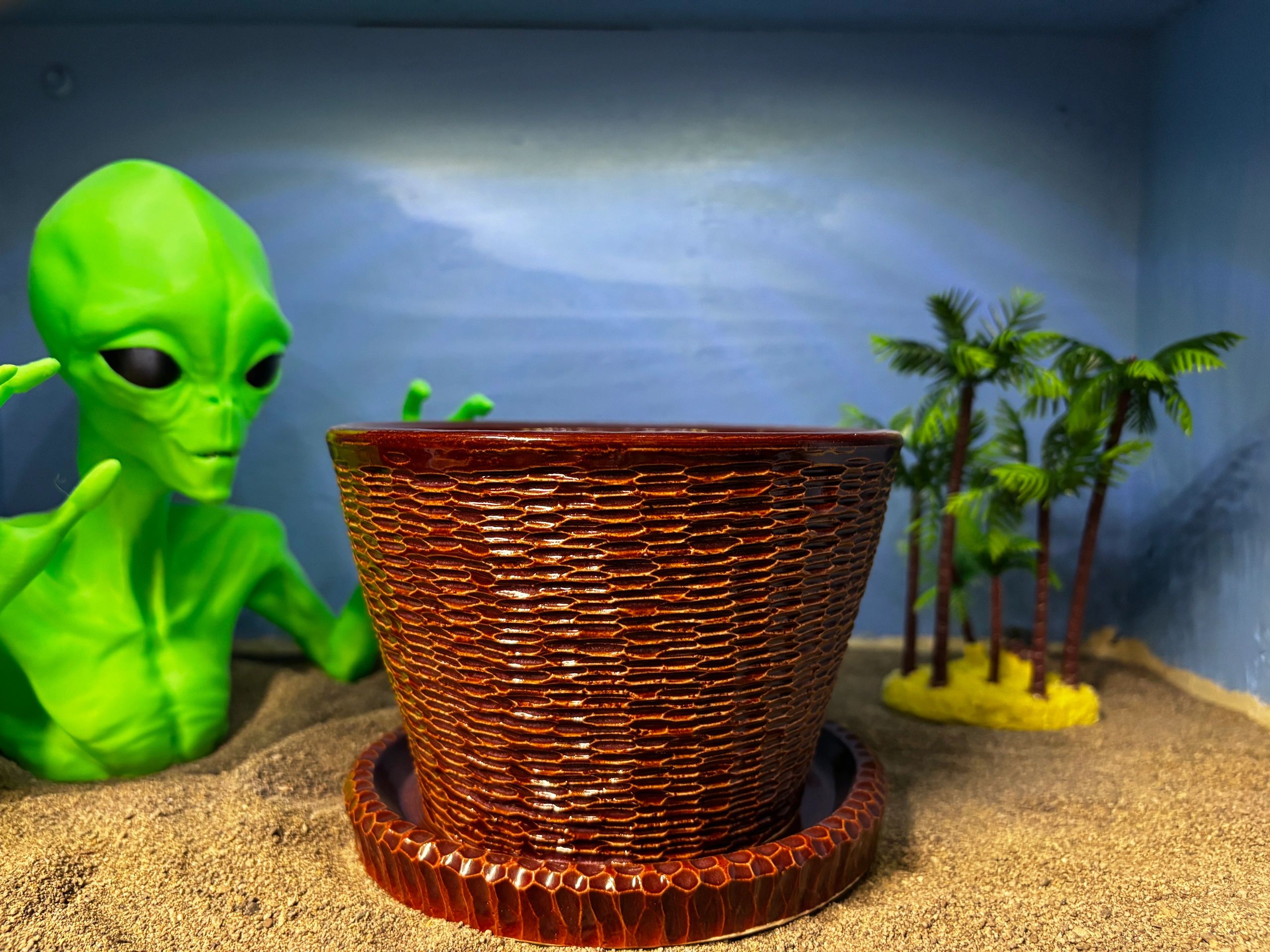 A green alien figure behind a brown textured pot on sandy surface with miniature palm trees.