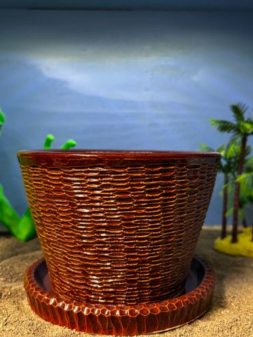 A green alien figure behind a brown textured pot on sandy surface with miniature palm trees.