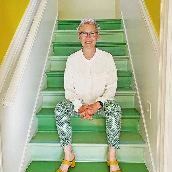 Laura in a white blouse, green pants, and mustard strappy shoes, sitting on painted green stairs
