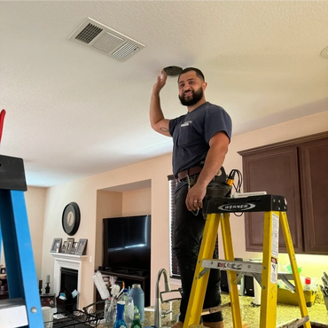 Technician fixing ceiling vent while standing on a yellow ladder in a kitchen.