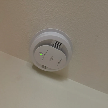 White smoke detector mounted on a ceiling with a green indicator light.