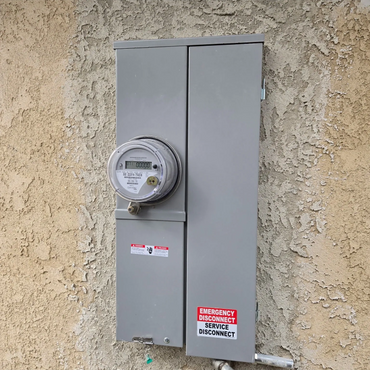Gray electrical meter box mounted on a textured wall with warning labels.