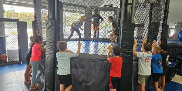 Kids watch a sparring match in a fenced gym area.