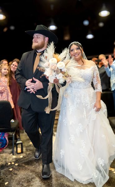 Newlyweds walking down the aisle, the bride in a lace dress and the groom in a black suit and cowboy hat.