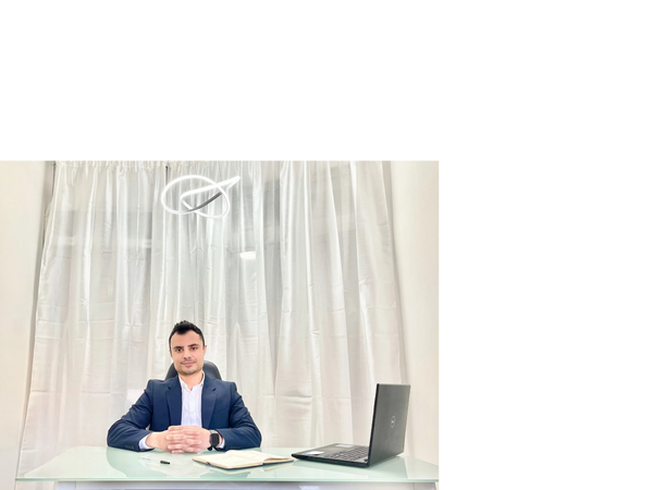 Man in a navy suit sitting at a glass desk with a laptop and notebook.